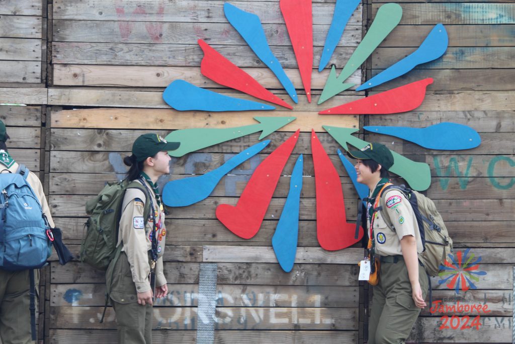 Two Japanese Scouts facing each other and smiling in front of Noa, the EIJ 2024 logo.