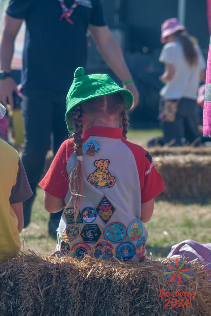 A rainbow wearing a frog bucket hat, sitting on a hay bale and facing away from the camera. Her back is covered in various badges. 