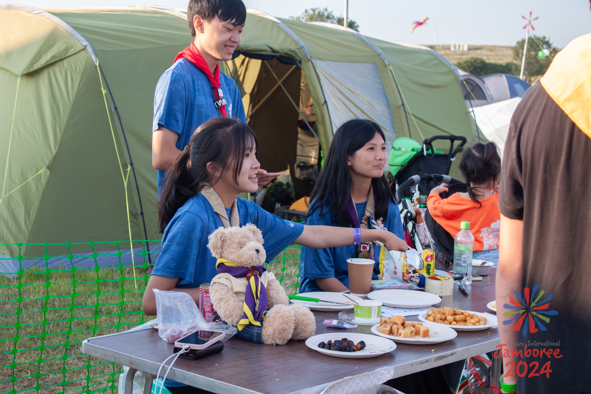 Some young people sitting behind a table covered in food. A teddy bear also sits on the table.