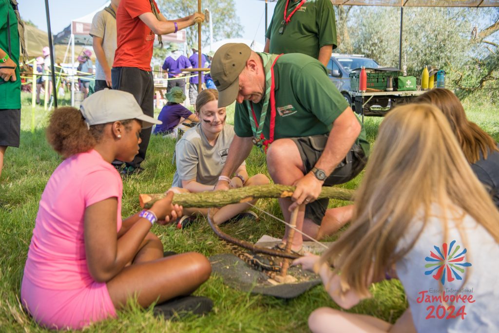 A leader holds a log in place while participants use a bow to start a fire through friction