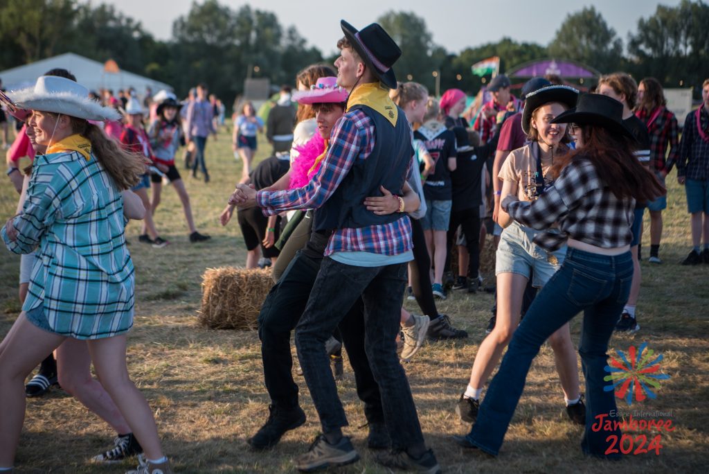 Participants in Wild West themed costume, dance in pairs, in hay bail-decorated field 