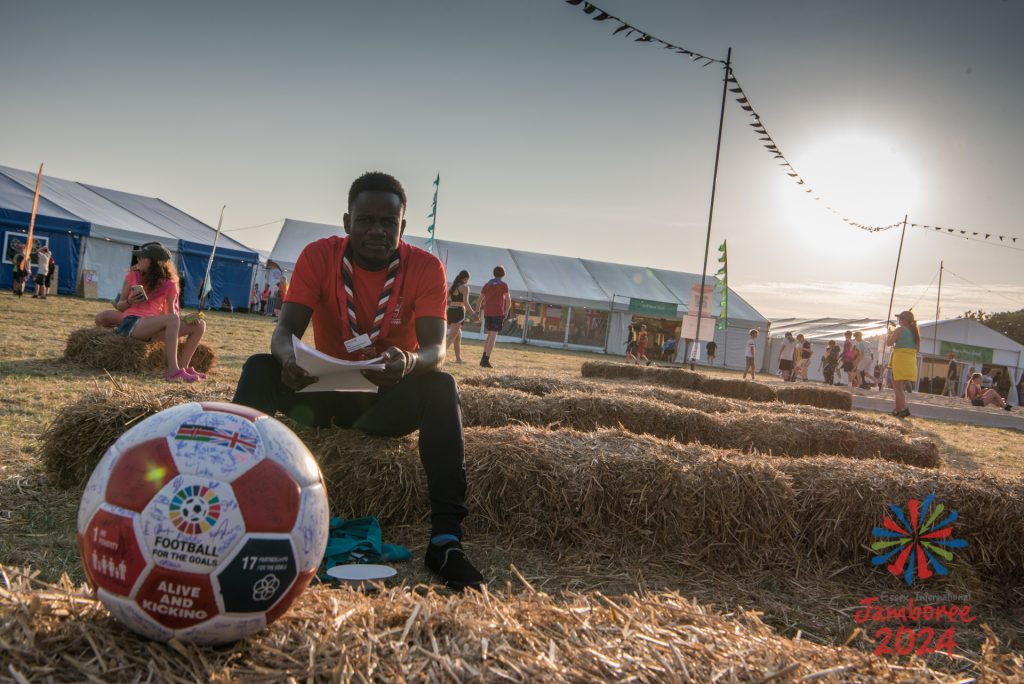 A football in the foreground, decorated with symbols of Global Development Goals, and signatures. Peter sits on hay bales at EIJ in the background.