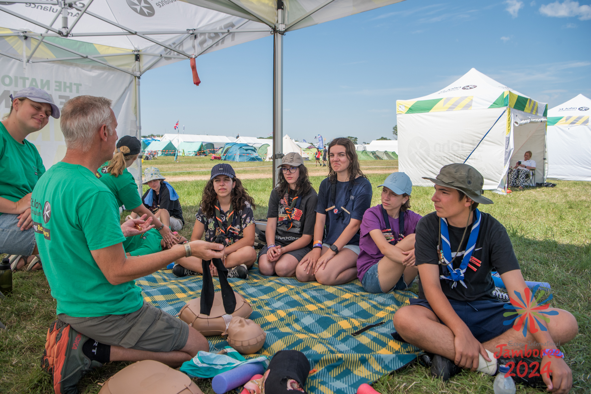 Participants sitting on a blanket listening to an instructor. A mannequin is laying in the middle of the blanket. A fake leg can be seen dangling from the ceiling.