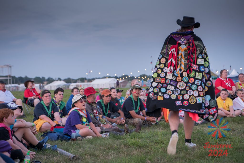 Participants enjoying the campfire, while the coordinator walks around singing. He's wearing his camp blanket as a cape, which is covered in badges and scarves. 