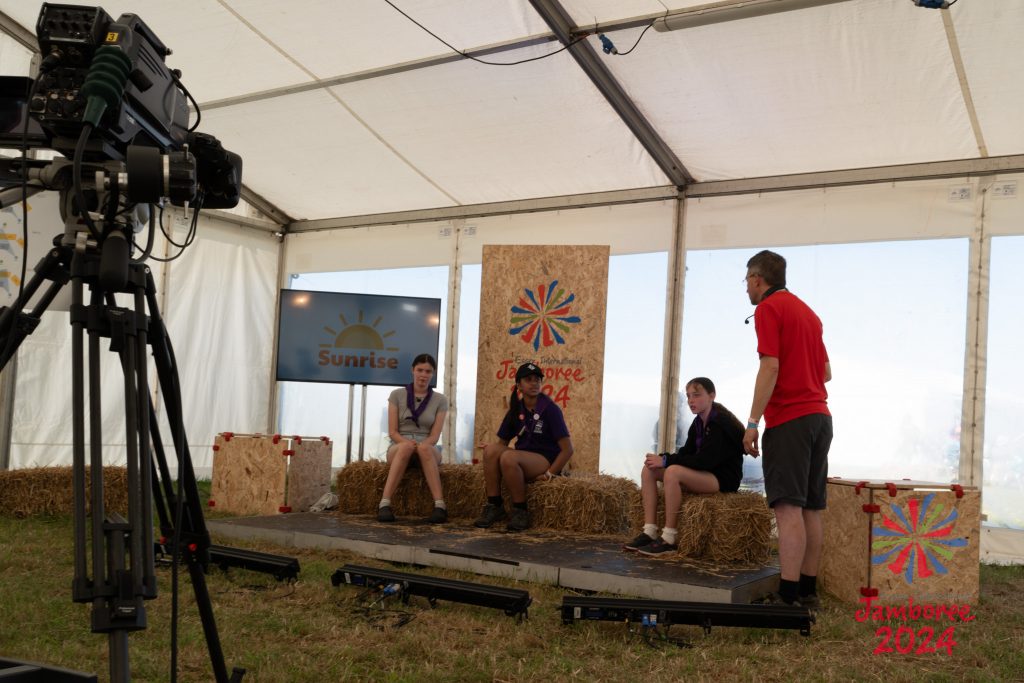 Three young people sitting on hay bales in front of a TV camera - about to present the news. A volunteer is standing to the side giving them instructions.