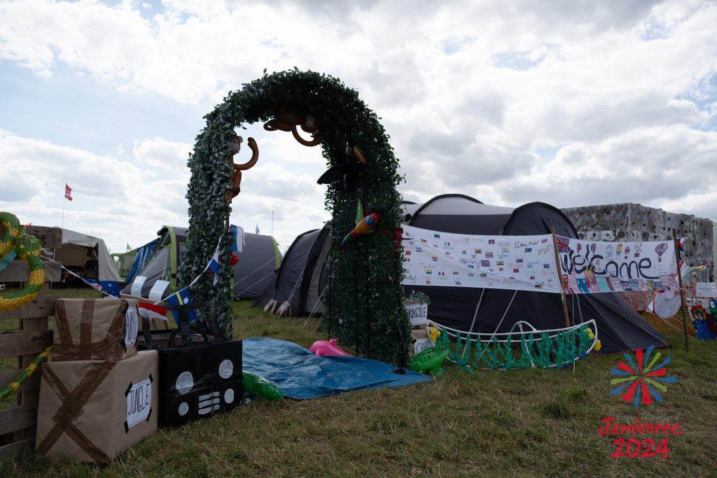 The gateway to a group's camp. The arch is covered in vines, with a water mat on the floor flowing under it. To the right there is a white banner decorated in flags, welcoming people to their camp.