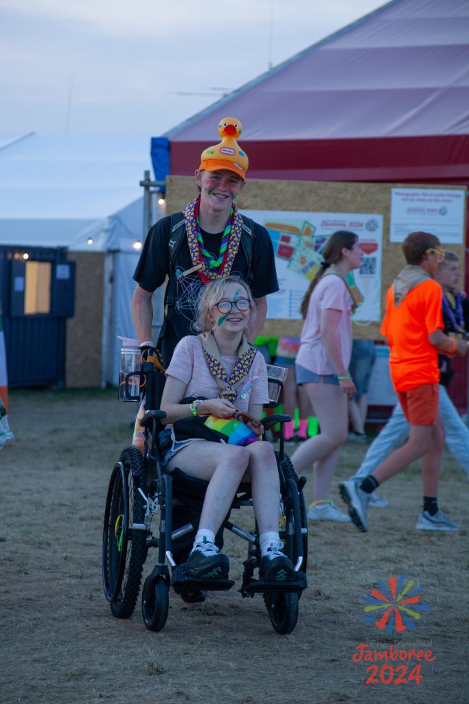 A young person in a wheelchair, being pushed by her friend, who is wearing a large rubber duck as a hat. Both are wearing duck-covered scarves and neon colours.