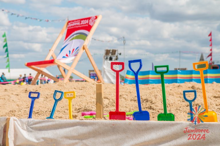 Different coloured spades stuck into the sand in a line. A large EIJ branded deckchair can be seen in the background.