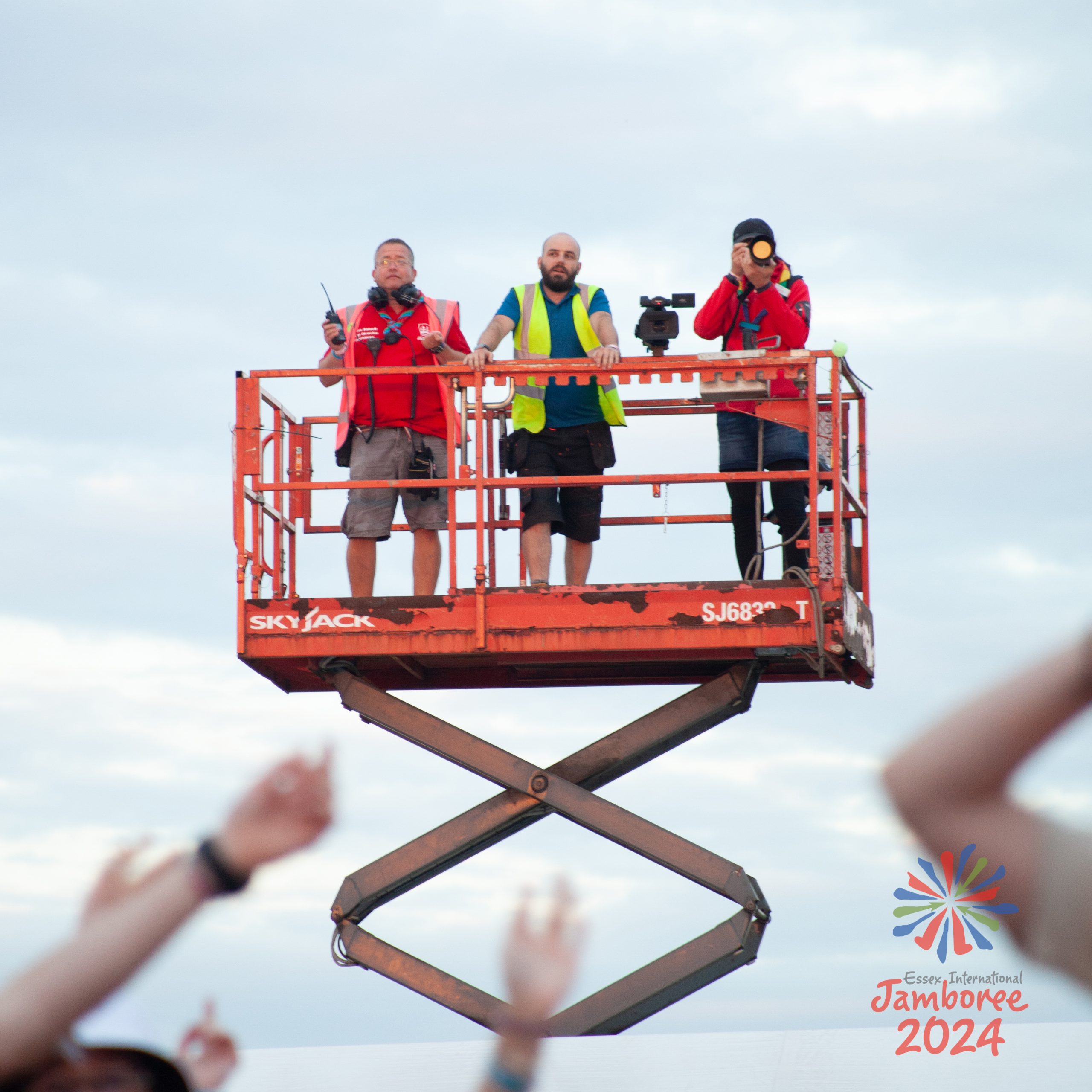 A scissor lift, extended high above the crowd, with volunteers wearing high-vis jackets standing inside it. One of them is pointing a camera directly towards the photographer.