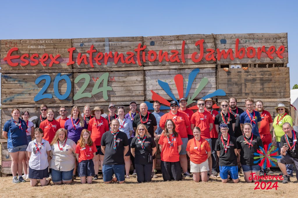 A group of people standing and kneeling in front of the Essex International Jamboree 2024 sign.