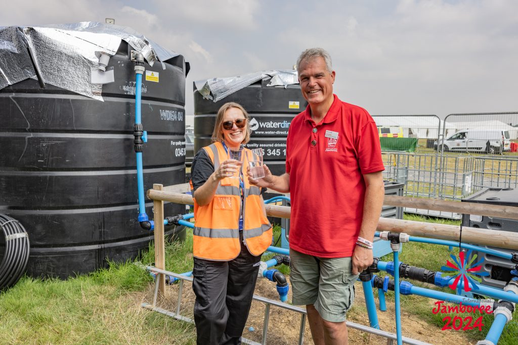 Two adults standing in front of two large water tanks, clinking glasses of water.