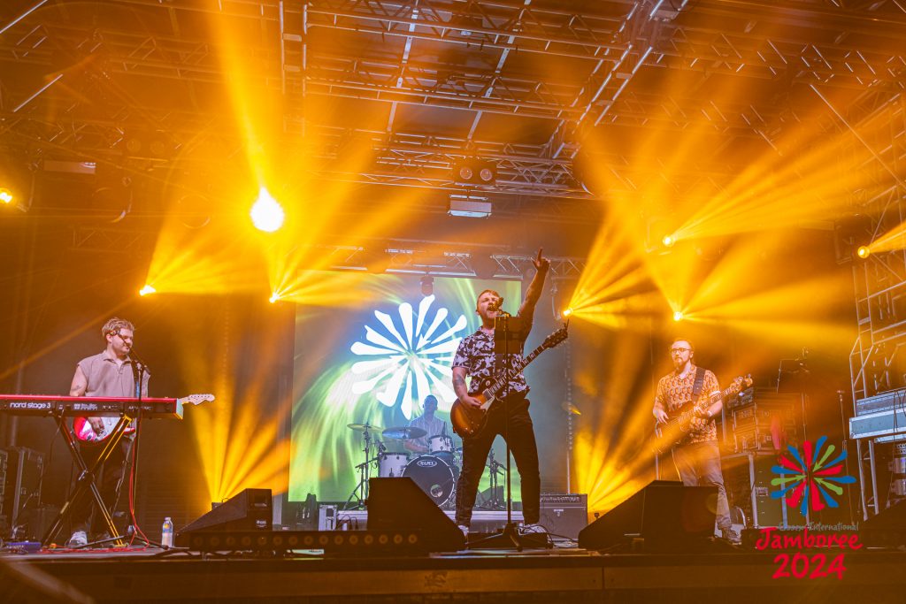 A band singing and playing on stage, with yellow lights streaking above. 